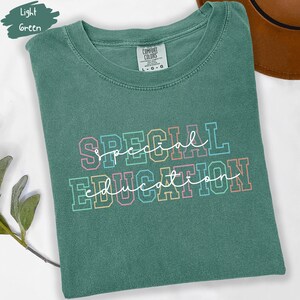 May include: A light green t-shirt with the words "SPECIAL EDUCATION" in a colorful, outlined font. The shirt is folded and has a tag that reads "COMFORT COLORS". The words are in a mix of script and block letters.