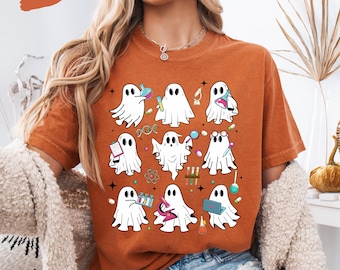Puede incluir: Camiseta naranja con ilustraciones de fantasmas blancos con accesorios científicos. Los fantasmas sostienen vasos de precipitados, tubos de ensayo y libros. La palabra "Yam" está en la esquina superior izquierda.
