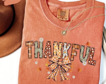 Könnte beinhalten: Ein korallenfarbenes T-Shirt mit dem Wort "THANKFUL" in einer Mischung aus Mustern, darunter Leopardenmuster und Karomuster. Unter dem Wort befindet sich eine große Schleife mit Leopardenmuster. Eine goldene Halskette mit Anhänger befindet sich auf dem Shirt.