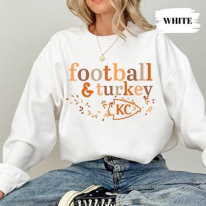 May include: A white crewneck sweatshirt featuring the text football & turkey in shades of orange and brown. Below the text is an orange outline of an arrowhead shape containing the letters KC, surrounded by small orange and brown leaf illustrations.