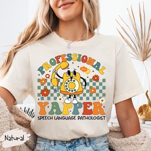 Puede incluir: Camiseta color crema con un diseño retro que presenta un personaje de teléfono amarillo. El texto dice "PROFESSIONAL YAPPER SPEECH LANGUAGE PATHOLOGIST" en una fuente lúdica, rodeado de flores y un fondo a cuadros.