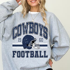 Puede incluir: Una sudadera gris claro de cuello redondo con un estampado gráfico azul oscuro de aspecto desgastado. El diseño incluye el texto COWBOYS y FOOTBALL, los números 19 y 60, y una ilustración central de un casco de fútbol americano azul.