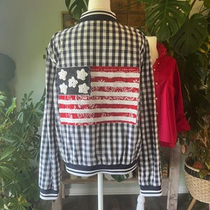 May include: A black and white gingham bomber jacket featuring a rectangular patch on the back. The patch displays a stylized American flag design created with red and white bandana fabric, white crocheted stripes, and white crocheted stars.
