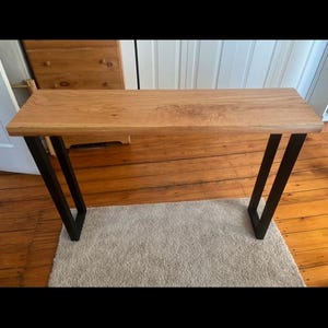 Puede incluir: Una mesa consola de madera con un acabado de madera natural y patas de metal negro. La mesa está sobre una alfombra beige.