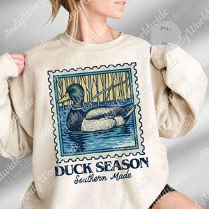 May include: Cream-colored sweatshirt featuring a postage stamp design with a duck in water and reeds. The stamp is outlined in blue with the words "DUCK SEASON Southern Made" below the image. A casual, comfortable top.