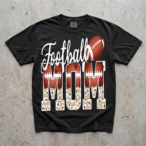 May include: A black short-sleeve t-shirt featuring the white cursive text Football above the block letters MOM. The letters are filled with a brown football texture, white laces, and a tan leopard print pattern, accented by small red hearts.