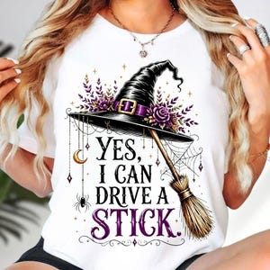 May include: A white t-shirt featuring a graphic of a black witch hat adorned with purple flowers and a gold buckle, a broomstick, a spider, a crescent moon, and a spiderweb. The text reads, "YES, I CAN DRIVE A STICK" in black and purple lettering.