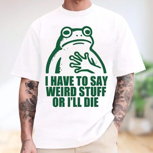 May include: A white short-sleeved t-shirt featuring a dark green graphic of a frog with its hand on its chest. Below the frog, the text reads: I HAVE TO SAY WEIRD STUFF OR I'LL DIE. The design is centered on the front of the garment.