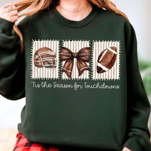 May include: A dark green long-sleeve sweatshirt featuring a graphic print of a brown football helmet, a brown bow, and a brown football, each inside a striped square. Below the graphics, the text reads, 'Tis the Season for Touchdowns'.