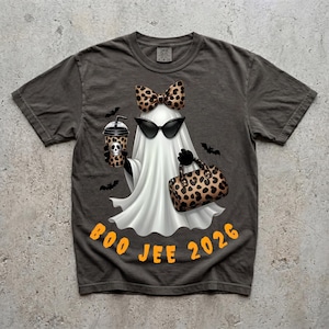 May include: A charcoal grey t-shirt featuring a white ghost graphic wearing a leopard print bow and black sunglasses. The ghost holds a leopard print cup with a skull and a matching leopard print handbag. The orange text reads BOO JEE 2026.