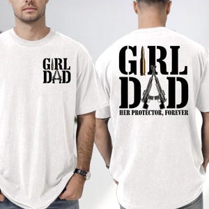 May include: White t-shirts with black text and graphics. One shirt has "GIRL DAD" printed on the front. The other shirt has "GIRL DAD" with two rifles and a bullet graphic on the back, with the text "HER PROTECTOR, FOREVER".