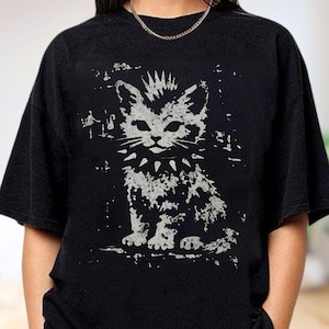 Puede incluir: Una camiseta negra de manga corta con un estampado gráfico gris claro desgastado de un gato con un collar de pinchos y un peinado mohicano. El gato aparece sentado y el diseño general tiene una apariencia texturizada y envejecida.