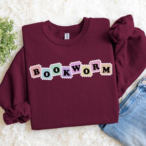 Peut inclure: Un sweat-shirt bordeaux avec le mot "BOOKWORM" imprimé sur la poitrine en lettres colorées, style timbre-poste. Le sweat-shirt est fait d'une matière douce et convient aux tenues décontractées. Le design est simple et attrayant.