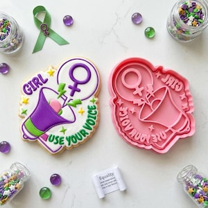 Puede incluir: Una galleta decorada y un cortador de galletas rosa con un diseño de megáfono y el texto "GIRL USE YOUR VOICE". La galleta tiene glaseado morado, verde, amarillo y blanco. También se muestra una cinta verde, chispas y pequeñas cuentas de vidrio.
