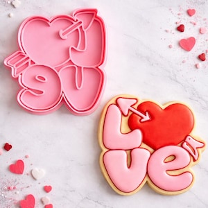 Op de afbeelding: Een roze plastic koekjesvorm en een versierde koek, beide met de tekst "LOVE" met een hart doorboord door een pijl. De koek is roze en rood met een gele rand. Hartvormige sprinkles maken het Valentijnsdagthema compleet.