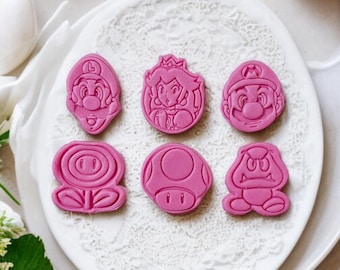 Puede incluir: Un plato blanco con seis cortadores de galletas rosas con forma de personajes de un videojuego popular. El plato está rodeado de galletas y huevos. Los cortadores de galletas incluyen a Mario, Luigi, la Princesa Peach y otras formas reconocibles.