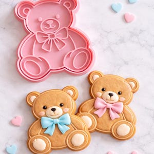 Può includere: Un tagliabiscotti rosa a forma di orsetto accanto a due biscotti a forma di orsetto decorati. I biscotti sono marroni con dettagli bianchi e hanno un fiocco. Un fiocco è blu, l'altro rosa. Ci sono decorazioni a forma di cuore rosa e blu sparse.