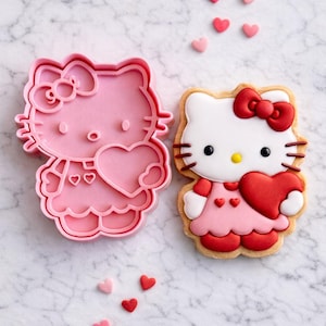 Puede incluir: Un cortador de galletas de plástico rosa y una galleta decorada con forma de gato de dibujos animados sosteniendo un corazón. La galleta tiene glaseado blanco, un lazo rojo y un corazón rojo. Pequeños adornos en forma de corazón están esparcidos.