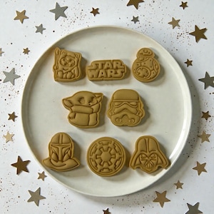 May include: A collection of Star Wars-themed cookie cutters in various character shapes, including Yoda, Darth Vader, and a Stormtrooper. The Star Wars logo is also present. The cutters are arranged on a white plate, surrounded by star-shaped decorations.
