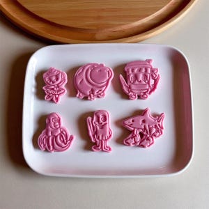 Könnte beinhalten: Rosa Ausstechformen in verschiedenen Formen, darunter eine Ballerina, ein Schwein, eine Stammesfigur, ein Oktopus, eine Strichmännchen und ein Hai. Die Formen liegen auf einem weißen, rechteckigen Teller.