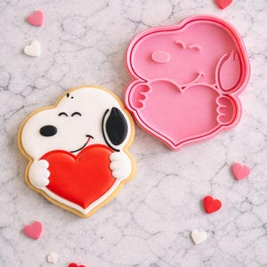 May include: A decorated biscuit and a pink cookie cutter shaped like a cartoon dog holding a red heart. The biscuit has white icing, a black ear, and a red heart. Small heart-shaped sweets are scattered around.