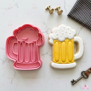 May include: A pink plastic beer mug cookie cutter next to a decorated beer mug biscuit. The biscuit is yellow with white foam and a white handle. Gold cufflinks and a key are also in the image.