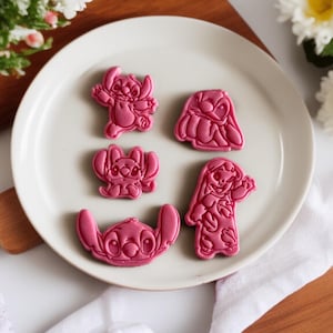 Może przedstawiać: Pięć różowych foremek do ciastek w kształcie postaci Stitch z filmu Lilo & Stitch, ułożonych na białym talerzu. Foremki przedstawiają Stitcha w różnych pozach i rozmiarach, idealne do pieczenia.