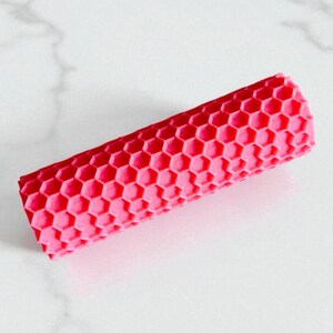 Puede incluir: Un objeto cilíndrico de color rosa brillante con un patrón de panal. El objeto es de un color sólido y parece estar hecho de un material flexible. El patrón de panal está espaciado uniformemente en toda la superficie.