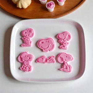 May include: A collection of pink Snoopy-shaped cookie cutters on a white rectangular plate. The cutters are in various poses, including standing, sitting, and lying down. The image also shows baked goods on a wooden tray.