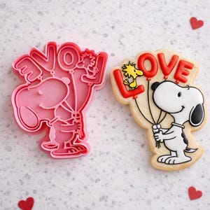 May include: A pink Snoopy cookie cutter and a decorated cookie featuring Snoopy holding balloons that spell "LOVE". The cookie is decorated with red and black details. Red heart-shaped confetti is scattered around the image.