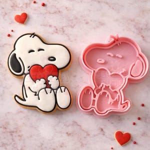 Puede incluir: Una galleta con forma de Snoopy sosteniendo un corazón rojo, junto a un cortador de galletas rosa de Snoopy. La galleta tiene contornos y detalles negros. Hay chispas rojas en forma de corazón y pequeños puntos rojos esparcidos. El fondo es un mármol rosa claro.