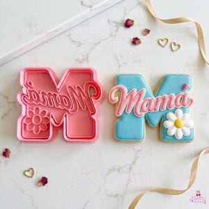 May include: A pink cookie cutter and a decorated cookie, both in the shape of the letter "M" with the word "Mama" in cursive. The cookie is light blue with pink frosting and a white daisy.