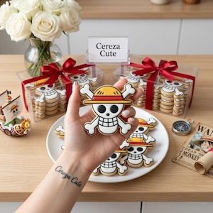 May include: A hand holds a skull and crossbones cookie with a yellow and red straw hat, matching the design of cookies in two clear boxes tied with red ribbons. A small ship figurine, a compass, and a sign reading Cereza Cute are visible.