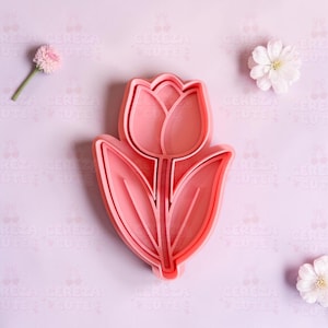 May include: A pink tulip-shaped cookie cutter with a detailed design of petals and leaves. The cutter is made of plastic and is designed for baking. The background is a light purple color with small white flowers and a pink flower.