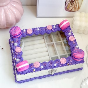 Puede incluir: Una caja de joyería púrpura y blanca con una tapa transparente. La caja está decorada con macarons rosas, piruletas y purpurina. Tiene un compartimento para anillos y una sección con varios compartimentos más pequeños.