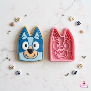May include: A decorated cookie and a pink cookie cutter shaped like a cartoon dog's face. The cookie is blue, white, and tan, with black details. The cutter is pink. Paw print and lavender decorations surround the cookie and cutter.