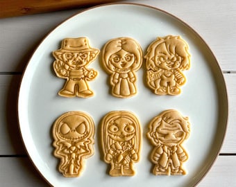May include: Six light brown cookies shaped like horror movie characters, including Freddy Krueger and Chucky, presented on a white plate with a brown rim. The cookies are intricately detailed and baked.