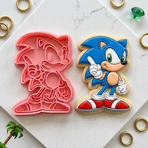 May include: A pink plastic cookie cutter shaped like Sonic the Hedgehog beside a decorated sugar cookie of Sonic. The cookie is blue, white, red, and tan. Gold rings and a green gem are scattered around the cookie and cutter.