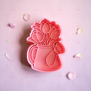 Może przedstawiać: Różowa, plastikowa wykrawaczka do ciastek w kształcie konewki z tulipanami. Wykrawaczka leży na różowej powierzchni z rozrzuconymi płatkami kwiatów. Tekst "CEREZA CUTE" powtarza się na krawędziach.