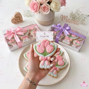 Könnte beinhalten: Ein Bouquet aus rosa Tulpenkeksen mit blauen und weißen Details, auf einem Teller präsentiert. Die Kekse sind mit rosa Schleifen verziert und in transparenten Boxen mit rosa und lila Bändern verpackt. Im Hintergrund steht eine Vase mit rosa und weißen Rosen.