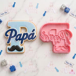 May include: A light blue cookie shaped like the letter "P" with the word "Papá" in dark blue script and a black mustache. Next to it is a pink cookie cutter in the same shape. The image includes small gift boxes and tie-shaped confetti.