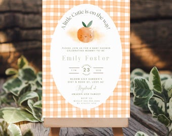 May include: A baby shower invitation on a small wooden easel. The invitation features an orange gingham background and an oval with the text "A little Cutie is on the way!" and details for Emily Foster's baby shower.