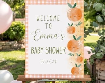 May include: A wooden easel displays a baby shower sign. The sign reads "Welcome to Emma's Baby Shower" with the date 07.22.25. Orange illustrations with smiling faces and green leaves decorate the border. The sign has a gingham border.