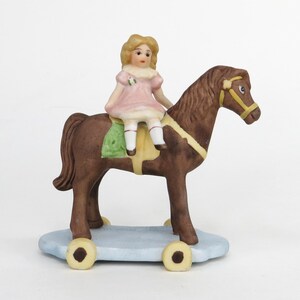 Könnte beinhalten: Eine Porzellanfigur eines Mädchens in einem rosa Kleid, das auf einem braunen Schaukelpferd mit gelben Rädern reitet.