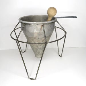 May include: A conical metal food mill or chinois strainer featuring a fine-mesh bottom, a sturdy wire stand with three legs, and a wooden pestle resting inside the basin. The kitchen tool has a metallic silver finish and a long, flat handle.