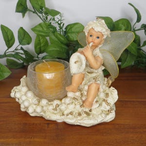 May include: A decorative candle holder featuring a cherubic angel figurine with translucent wings. The angel sits on a cloud-like base, holding a calla lily. A yellow candle sits in a crackled glass holder.
