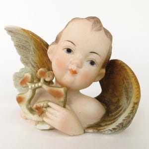 Könnte beinhalten: Eine Porzellanfigur eines Cherubins mit Flügeln und einer Harfe. Der Cherub hat hellbraunes Haar, blaue Augen und einen leichten rosa Rouge auf den Wangen. Die Harfe ist in Braun- und Grüntönen bemalt.