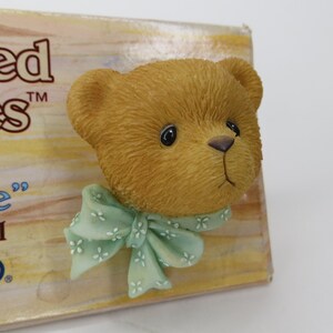 May include: A sculpted tan bear head figurine featuring a light green bow with a small white floral pattern around the neck. The bear head is attached to a rectangular cardboard backing with partial text visible on the left side including the letters ed and S.