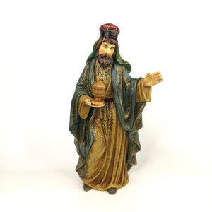 May include: A figurine of a wise man, part of a nativity scene. The figure wears a green and gold robe and a red crown. He holds a golden lantern in one hand and gestures with the other. The figurine is on a white background.