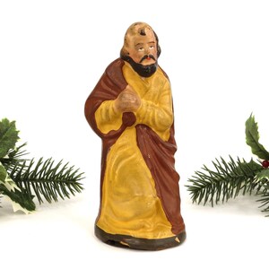Può includere: Statuina dipinta a mano di un uomo in una lunga veste, con una sottoveste gialla e una sopravveste marrone. La figura ha la barba e sta in piedi con le mani giunte. Agrifoglio e rami di pino sullo sfondo.